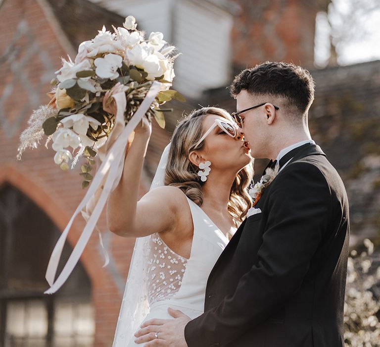 Bride in fitted wedding dress with mesh back and dot design wedding dress and sunglasses kisses groom in black tie also wearing sunglasses and neutral bouquet with ribbons