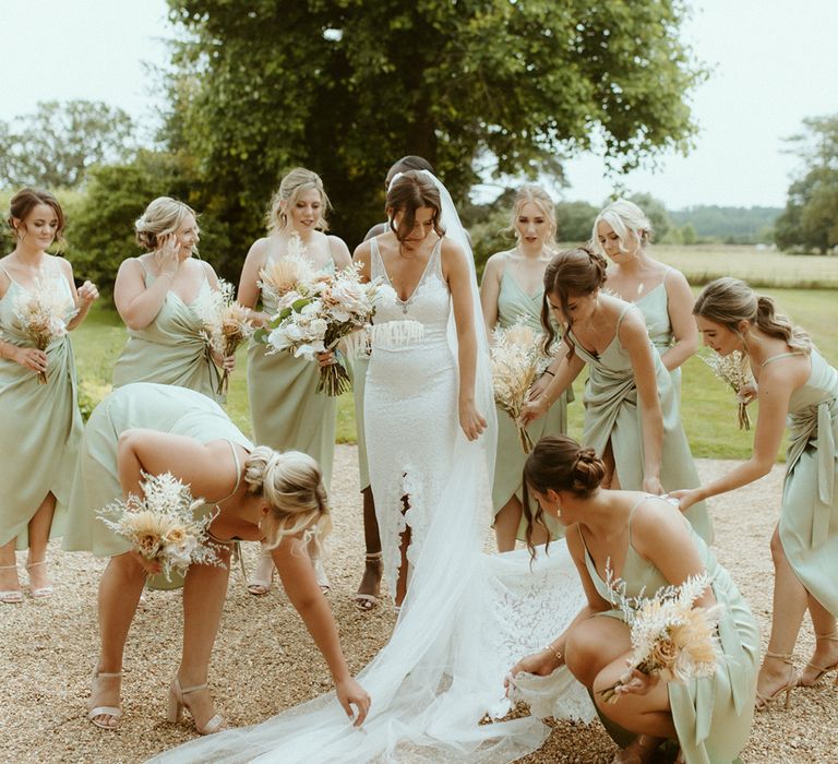 Bride in lace wedding dress with front slit and v-neck with bridesmaids in sage green dresses helping lay out the veil 