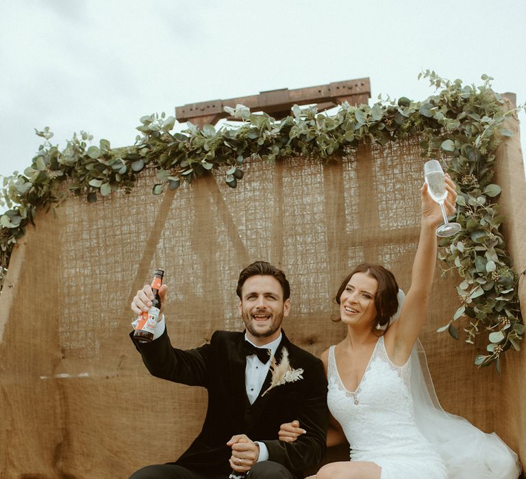 Groom in black tie lifts his drink as the bride does the same wearing white wedding heels with pearl anklet charm on back of tractor