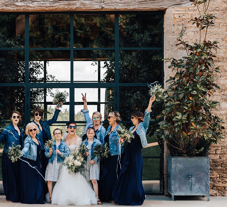 Bride poses with her bridal party all in blue and denim jackets with sunglasses for her rustic luxe wedding 