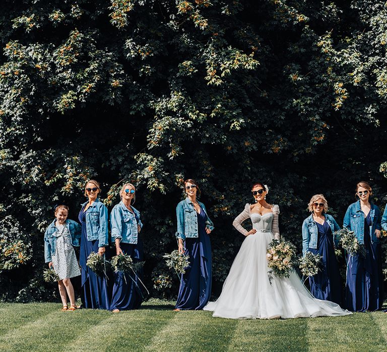 Bridesmaids in navy blue dresses wear denim jackets with sunglasses and pose with bride in sunglasses with red hair and hair bow 