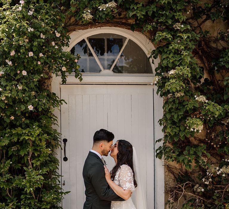 Bride & groom kiss in front of white door outdoors surrounded by green foliage at Northbrook Park