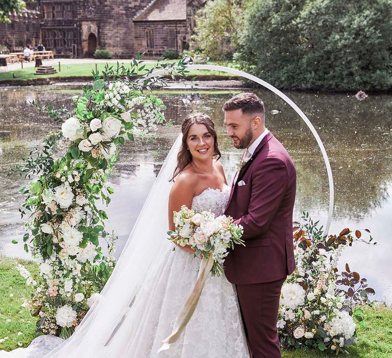 Bride in lace wedding dress and groom in a burgundy coloured suit standing in front of a hoop floral arch at East Riddlesden Hall