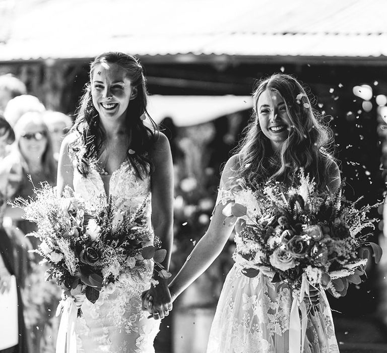 Brides with interlocked fingers walk together through confetti 