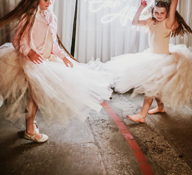 Flower girls wearing ballerina dresses and floral crowns dance in front of neon wedding sign