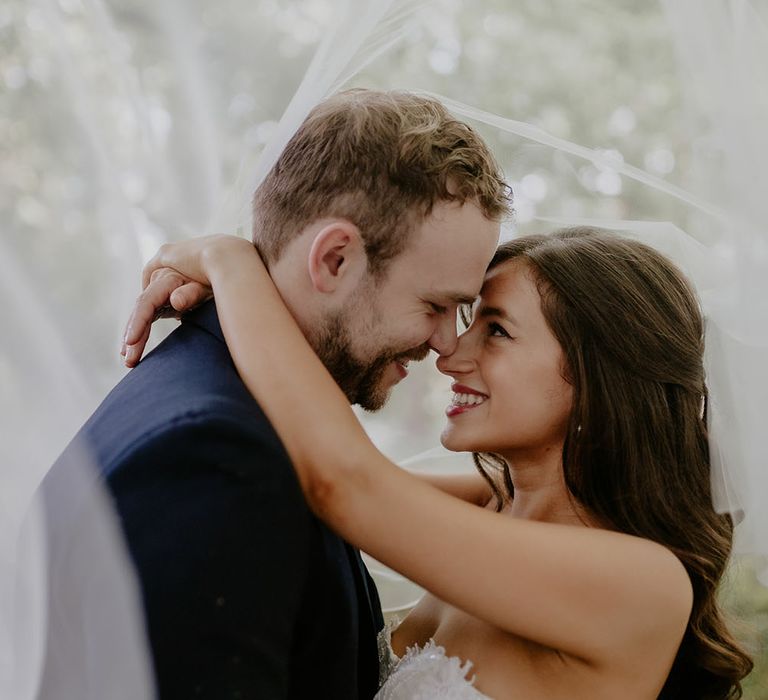 Bride & groom stand beneath veil as bride wears her brown hair in loose waves 