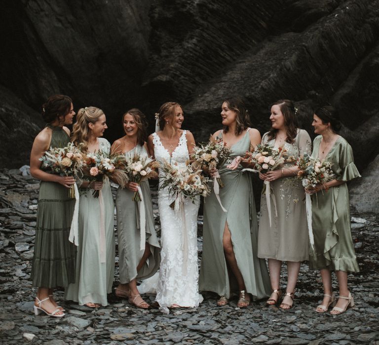 Bride stands with her bridesmaids who wear different shades of green bridesmaid dresses in differing styles as they hold champagne and rust coloured bouquets