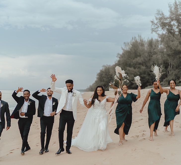 Bride & groom walk along the beach with groomsmen in black tie and bridesmaids in different styled green satin dresses 