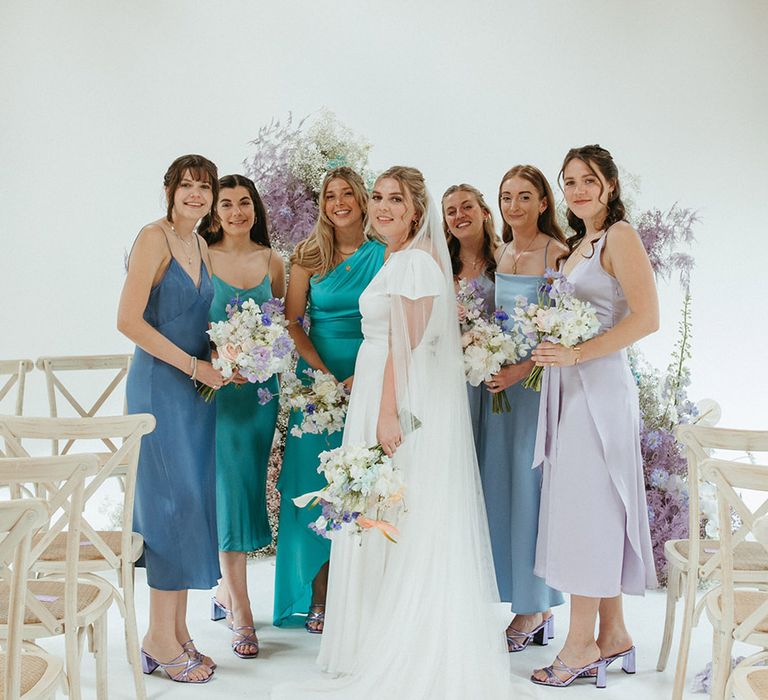 Bride stands with bridesmaids in different shades of blue bridesmaid dresses and matching purple metallic shoes 