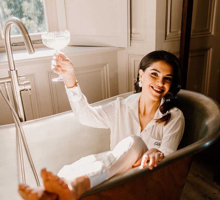 Bride lays out in a copper bath drinking champagne in white bridal pyjamas 