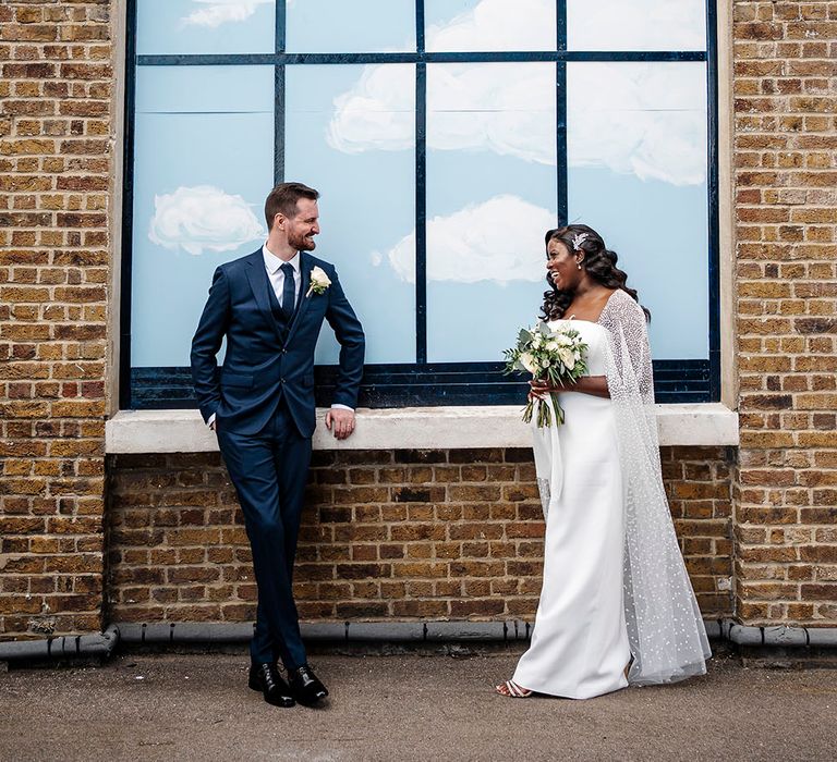 Bride & groom stand in front of painted sky window as bride wears glitter polka dot tulle wedding cape 