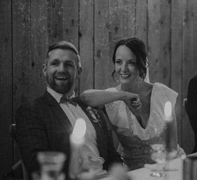 Bride and groom sit smiling and laughing as they listen to the wedding speeches 