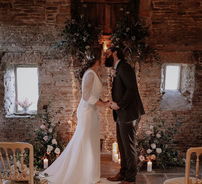 Bride and groom share their first kiss as a married couple with white and pink roses, candlelight and fairy lights for a rustic winter wedding 