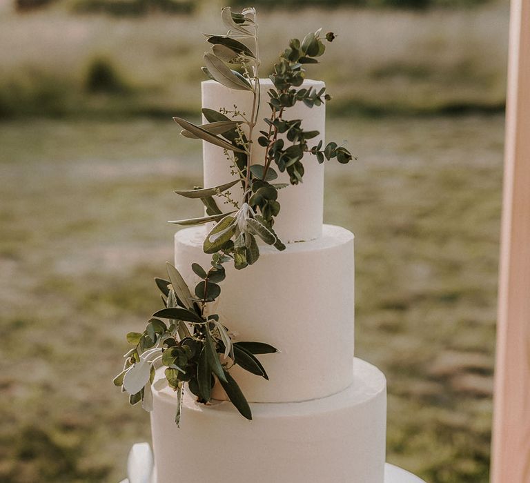 Three tier iced white wedding cake on a white and mint green cake stand decorated with greenery 