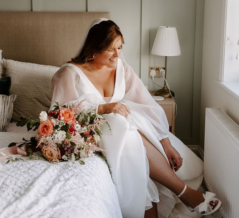 Bride wears full sheer sleeved romantic wedding dress whilst putting on strappy white bridal shoes with bow 