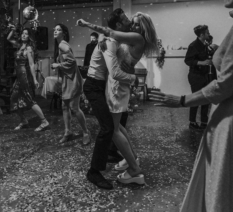 Bride in short satin dress with feather trimmed neckline and groom in dark tux kissing and dancing at Hackney Studios