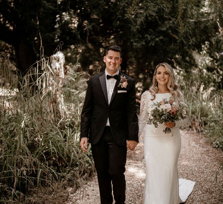 Groom in black tie walking with the bride in a Badgley Mischka wedding dress with soft pink bouquet and buttonhole 