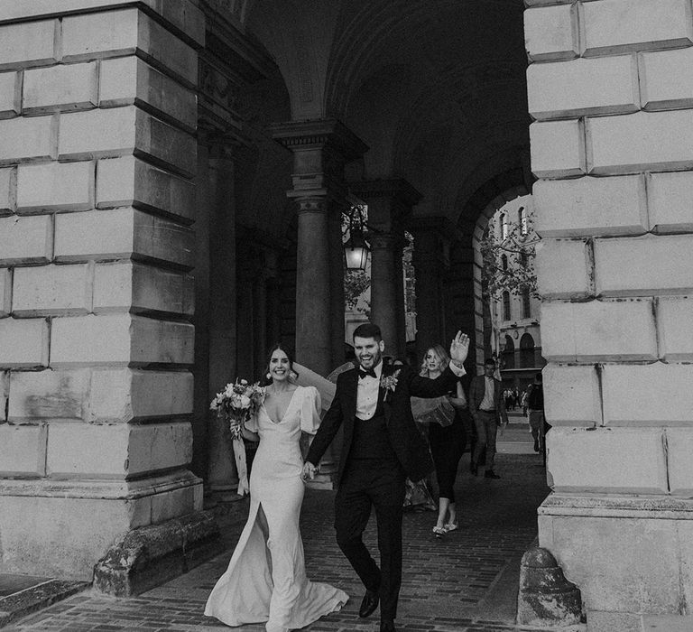 Bride in v neck front slit wedding dress holding neutral toned bouquet with groom in dark tux walking out of large archway at Somerset House