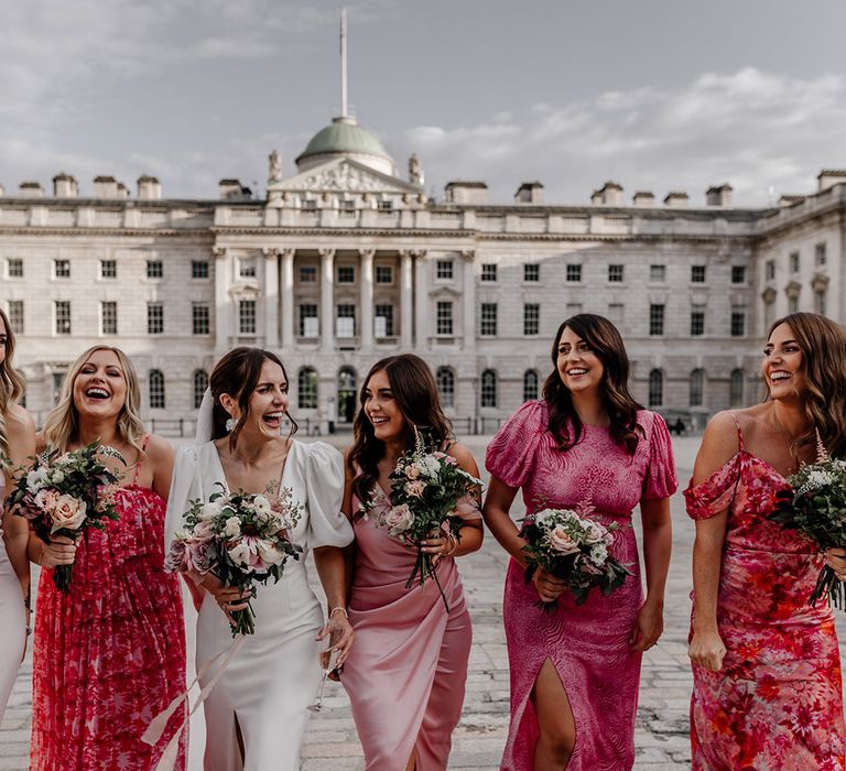 Bride in modern front slit wedding dress with puff sleeves arm in arm with bridal party wearing mismatched pink bridesmaid dresses and matching neutral bouquets