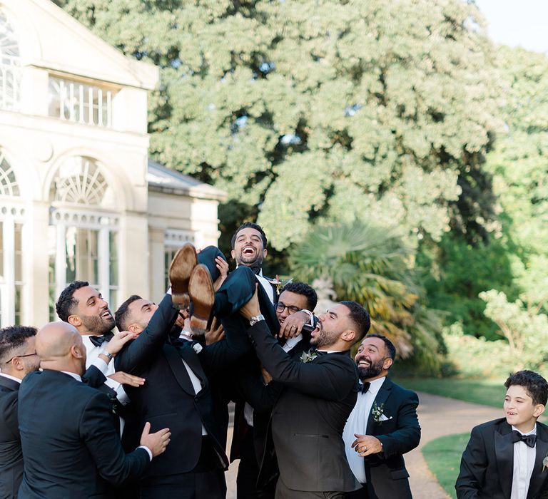 Groomsmen in black-tie lift groom in the air outdoors 