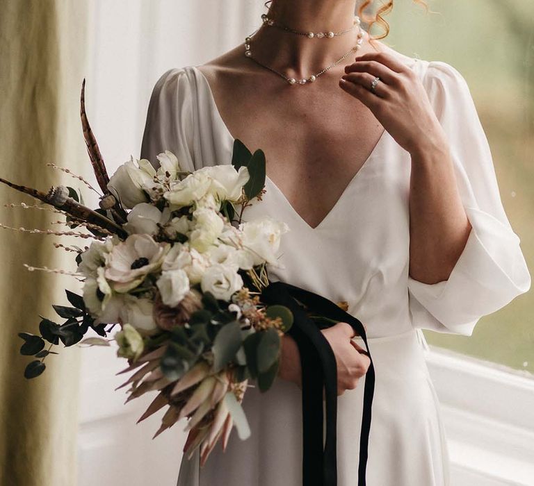 Bride in v neck satin wedding gown with double layered pearl necklace and holding minimalistic, neutral floral bouquet