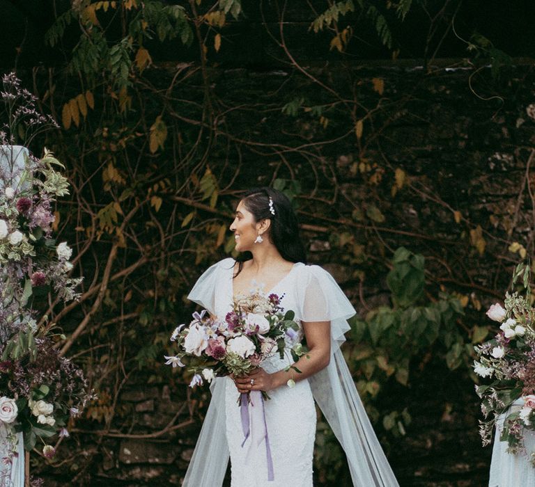 Bride in lace wedding dress with puff lace sleeves and bridal wings holding mixed floral bouquet with with white carnations, purple tulips, roses and foliage