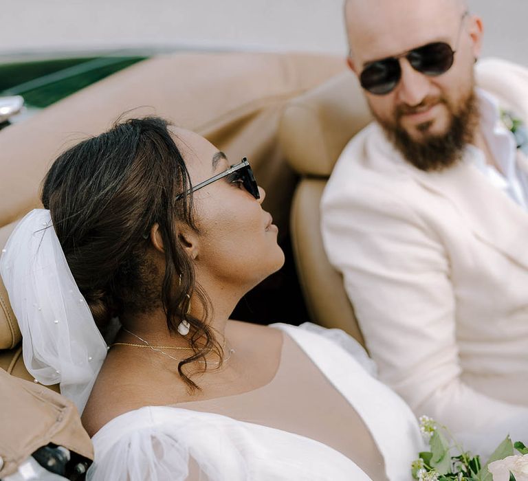 Bride and Groom look lovingly at each other whilst wearing sunglasses in a vintage Alfa Romeo Spidee