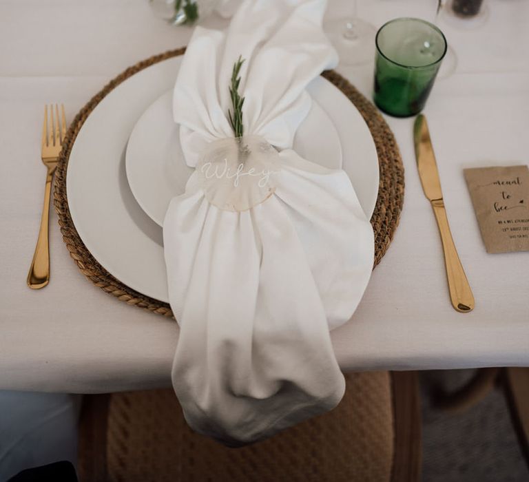 Place setting with wicker place mat, white plates, green coloured glassware, gold cutlery, with white napkin and frosted place name 