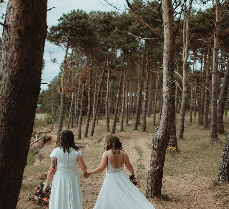 Brides walk together hand in hand as they explore the grounds of the venue in a woodland and beach 