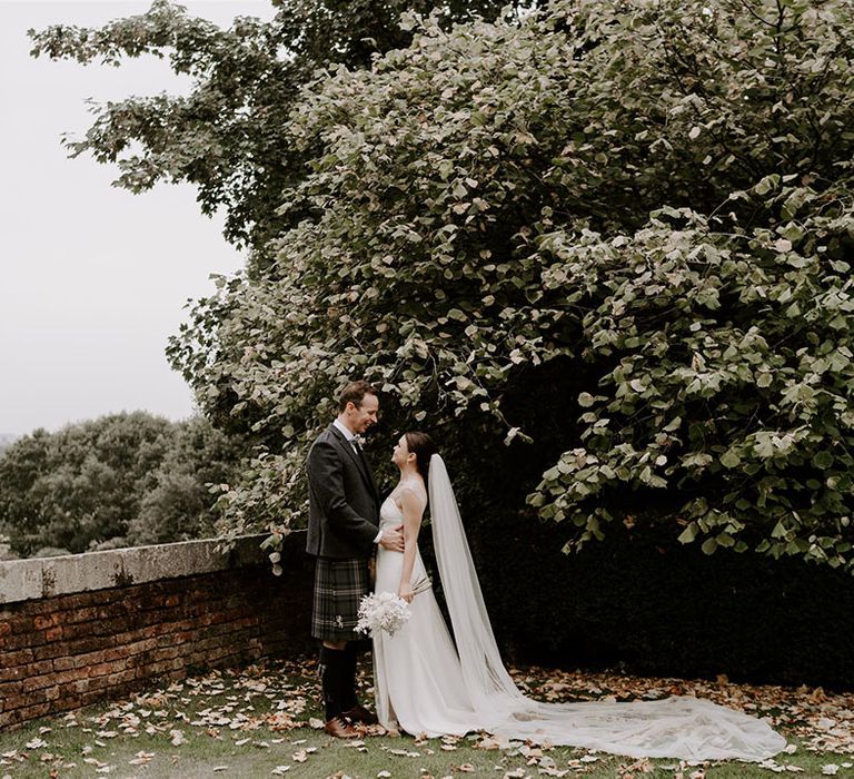 Groom in a kilt smiles lovingly at the bride in a sparkly wedding dress and cathedral length veil 