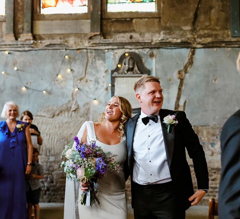 Bride holds colourful bouquet and stands beside her groom in black tie and floral buttonhole