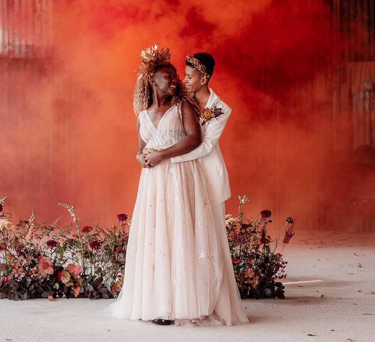 Bride in elegant white silk suit with autumnal dried floral boutonniere embracing bride in in sleeveless floaty peach gown and golden leaf crown with bright orange smoke bomb and dried floral arrangements 