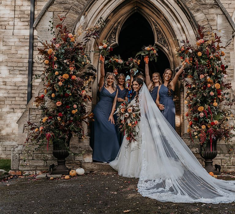 Bridesmaids in navy blue dresses posing with the bride in a lace wedding dress next to autumnal wedding flower columns with orange flowers