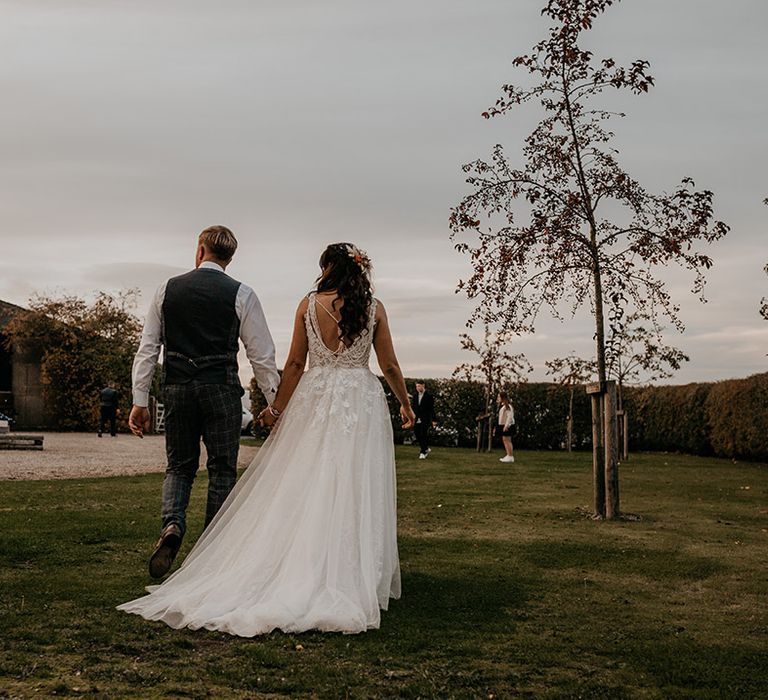 Groom in a grey waistcoat and the bride walk holding hands for couple portraits at the rustic barn wedding venue 