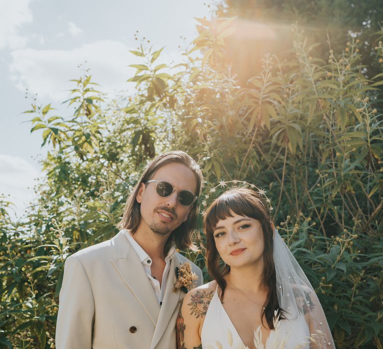 Bride in v neck ASOS wedding suit with gold halo headband and dried floral bouquet standing and smiling with groom in neutral-toned double-breasted ASOS suit and sunglasses