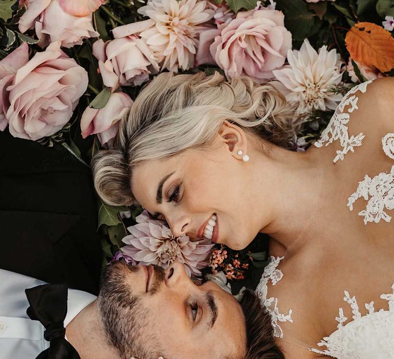 Groom in black tuxedo blazer with dried boutonniere and black bowtie lying on a bed of blush dahlias and garden roses with bride in  lace wedding dress with sheer, lace top and clusters of lace flowers on the shoulders and plunge neckline 