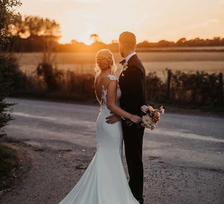 Bride in lace wedding dress with sheer, lace top and clusters of lace flowers on the shoulders and plunge neckline with button back details holding blush pink bridal bouquet embracing groom in classic black tuxedo looking at the sunset 