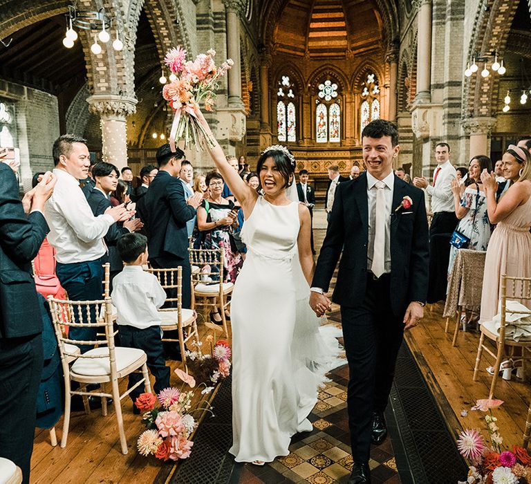 Bride wearing fitted wedding dress lifts her colourful bouquet in the air alongside her groom 