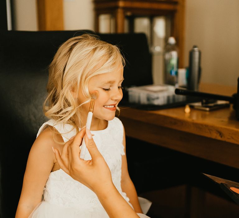 Blonde flower girl has blusher applied to her cheeks before wedding ceremony