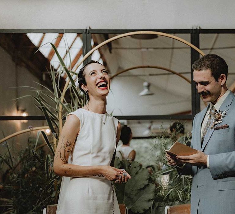 Lisa in fitted pre-loved wedding dress with bateau neckline laughs as Gabriel reads vows during non-denominational wedding ceremony