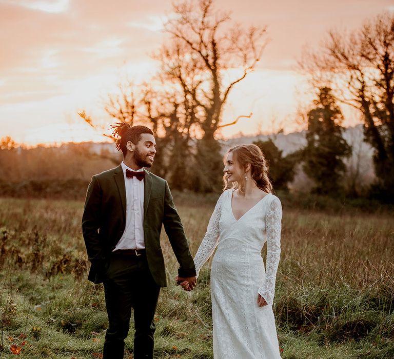 Bride in long sleeve lace wedding dress and groom in black tux with velvet burgundy bowtie holding hands at golden hour 