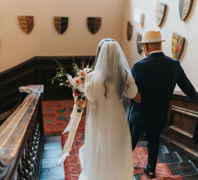 Father of the bride in navy suit with hat walking the bride down the stairs at Askham Hall for the relaxed intimate wedding 