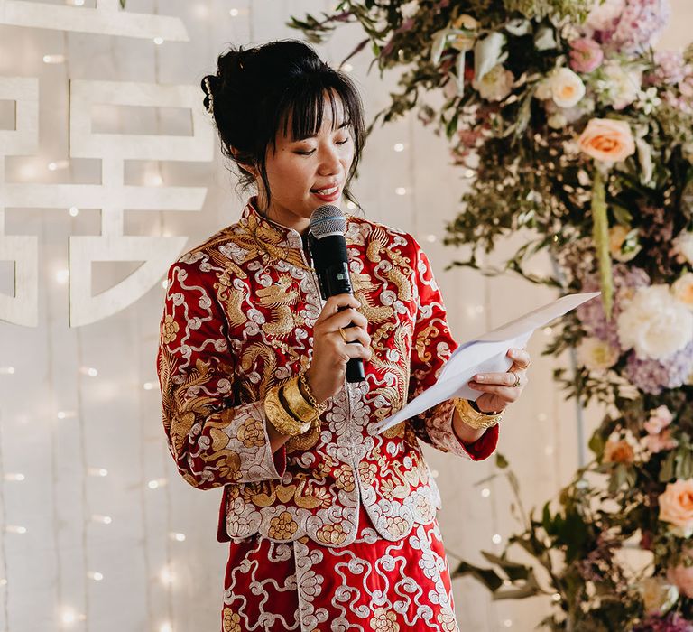 Bride in traditional red Chinese wedding dress with gold embellishment reads speech during ceremony 