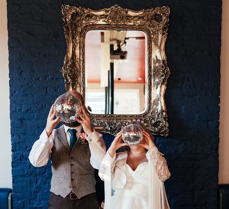 Groom in grey waistcoat, deep green tie and rose boutonniere standing with bride in midi-length bridal gown with lace embroidered sleeves and polka dot wedding boots holding up disco ball wedding decorations 