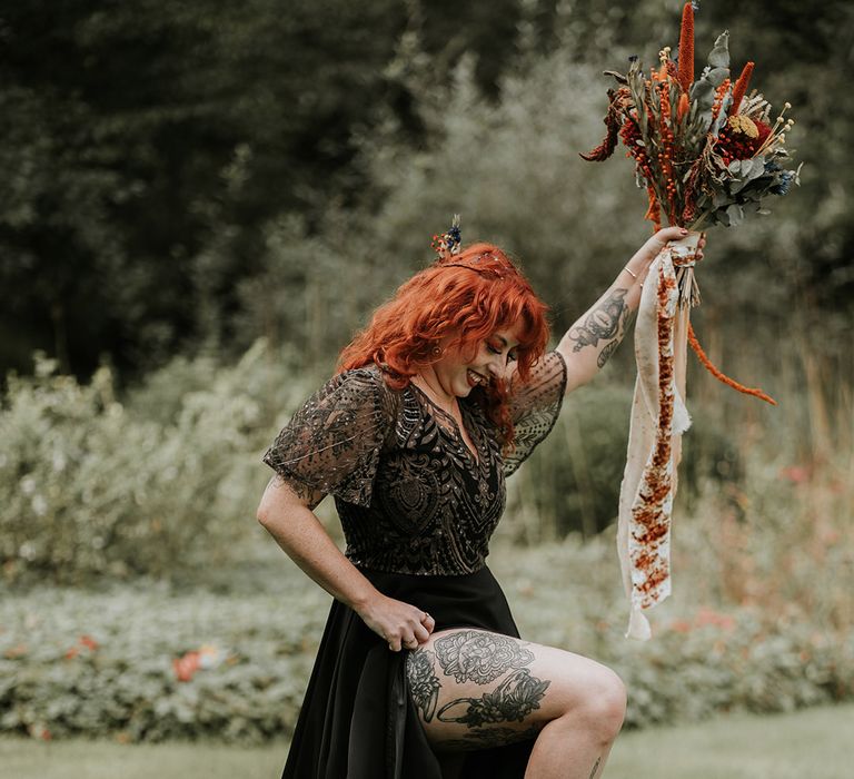 Bride in embellished v-neck short sleeve black wedding dress showing off her tattoos holding large burnt orange and eucalyptus dried flower bouquet with orange and off-white ribbons