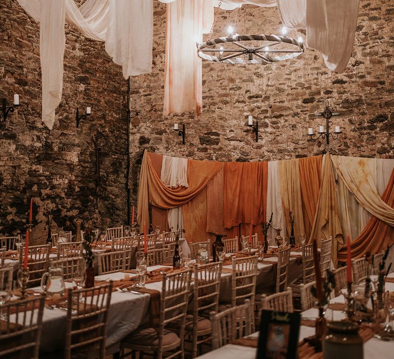 Rustic boho wedding reception room with festoon chandelier lighting, off-white and burnt orange wedding drapes decorations, fairy lights and dried flower centrepieces 