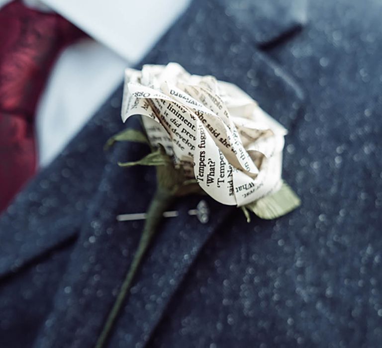 The groom in a dark blue suit with a red tie wearing a paper rose buttonhole made from the pages of the couple's favourite book 