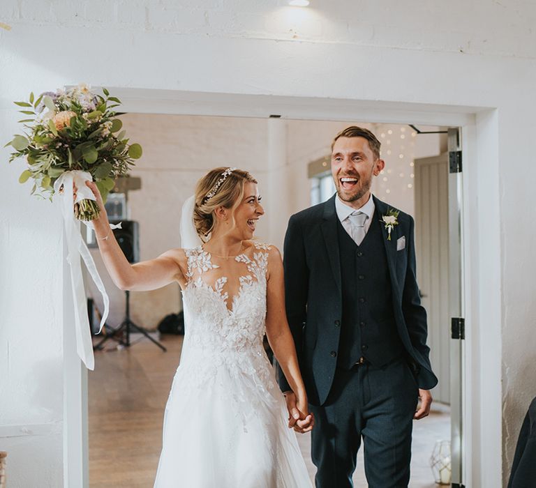 Bride in illusion lace Dando London wedding dress enters the wedding reception with the groom in a navy suit 