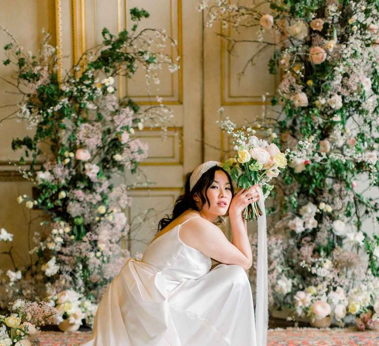 Bride in satin wedding dress with dramatic bow accent on the back posing with large rose, baby's-breath, foliage and dried flower floral columns holding classic spring bouquet 