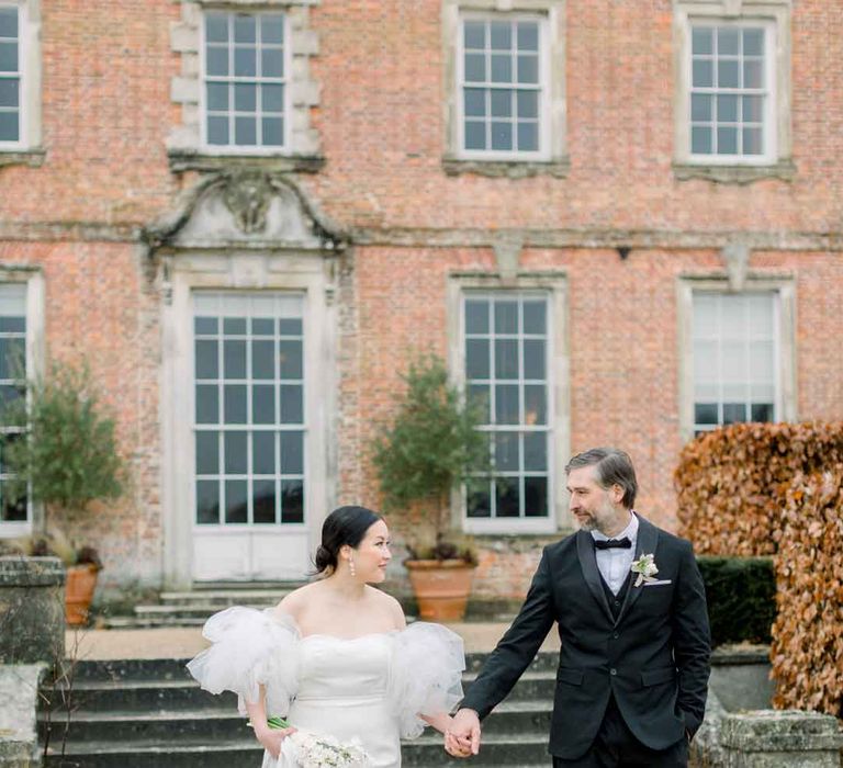 Bride in strapless wedding dress with puff tulle sleeves walking with groom in classic black tuxedo, black bowtie and boutonniere walking the grounds of St Giles House